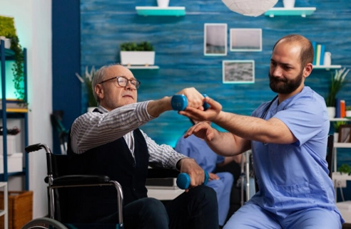 Therapist assisting elderly man in wheelchair with arm exercises using dumbbells for muscular dystrophy rehabilitation